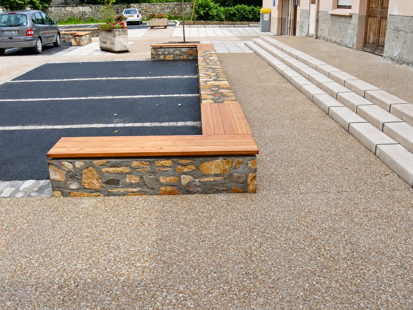 Un banc en maçonnerie de pierre surmonté d'une assise en bois longe un parking en enrobé et un sol en béton désactivé, près d'un escalier en pierre claire.
