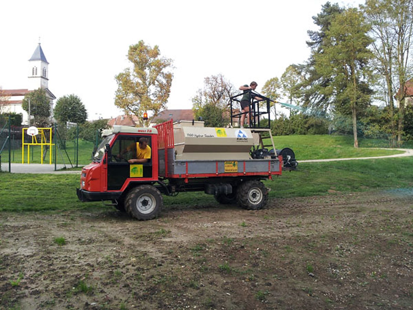 Un petit camion rouge équipé d'une cuve d'hydroseeding projette un mélange sur un terrain de jeux, avec deux personnes à bord et une église en arrière-plan.