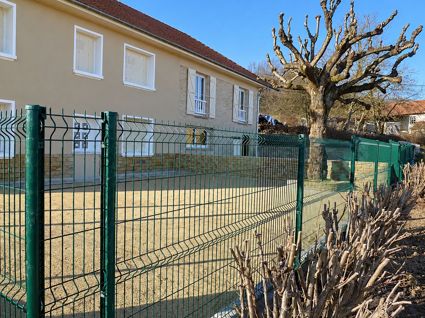 Une clôture en treillis métallique vert entoure une cour gravillonnée devant une maison beige à volets, avec un grand arbre taillé en arrière-plan.
