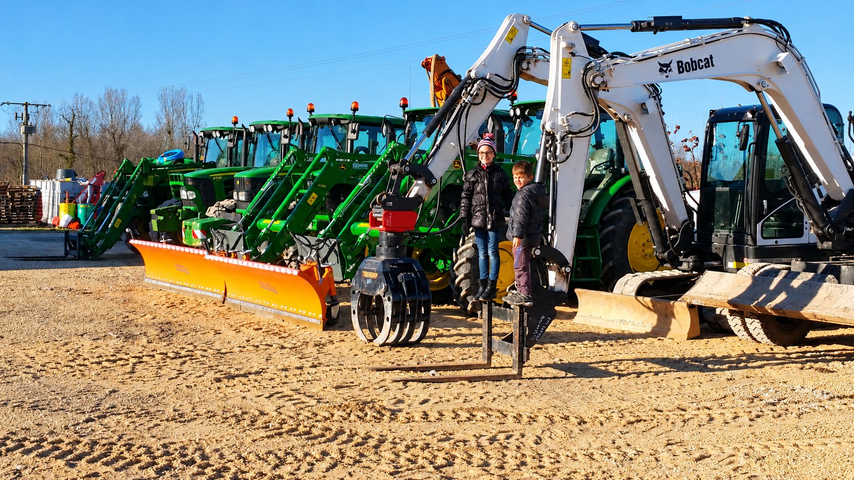 Deux enfants posent sur un terrain de sable devant une rangée de tracteurs verts équipés de lames à neige orange et de mini-pelles Bobcat blanches.