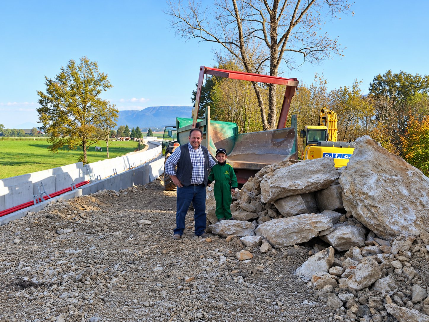 Un homme et un jeune garçon en combinaison verte posent sur un chantier de terrassement, à côté d'un tas de gros rochers, avec une pelleteuse jaune et un camion à benne en arrière-plan.