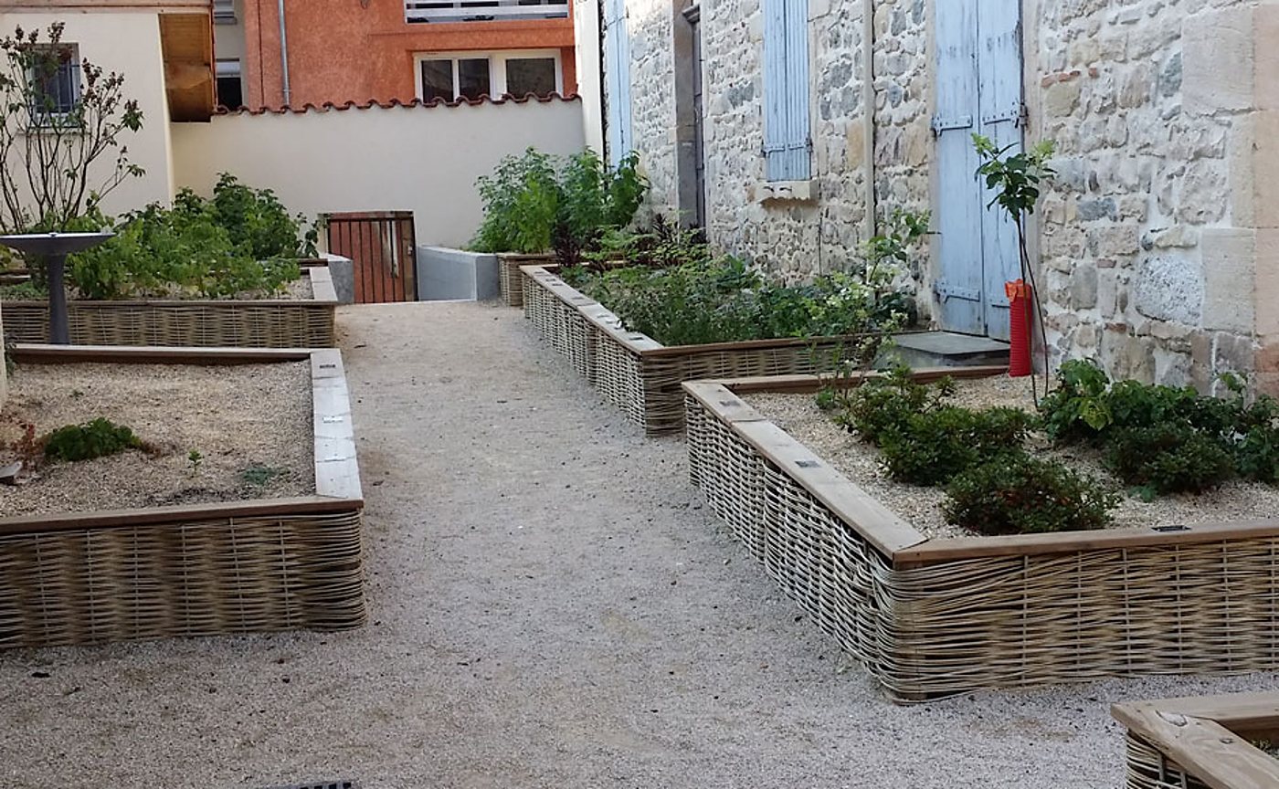 Des carrés potagers surélevés bordés d'osier tressé et de bois bordent une allée de gravier devant une façade en pierre aux volets bleus.