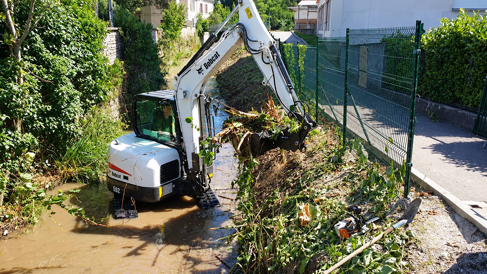 Mini-pelle Bobcat curant un cours d'eau bordé de végétation, godet chargé de branches et de racines.