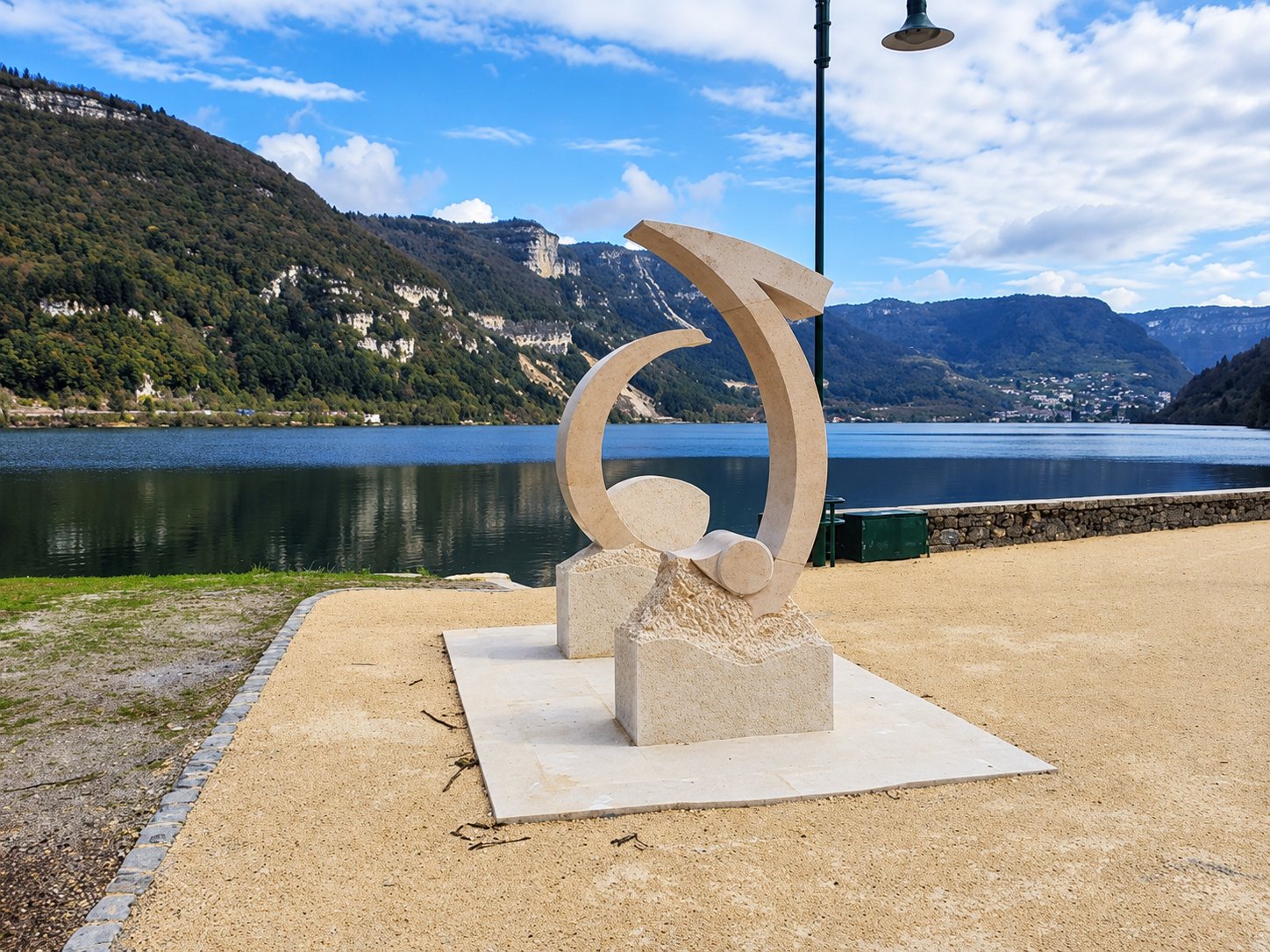 Une sculpture moderne en pierre claire en forme de croissant, posée sur un socle dallé au bord d'un lac entouré de collines boisées.