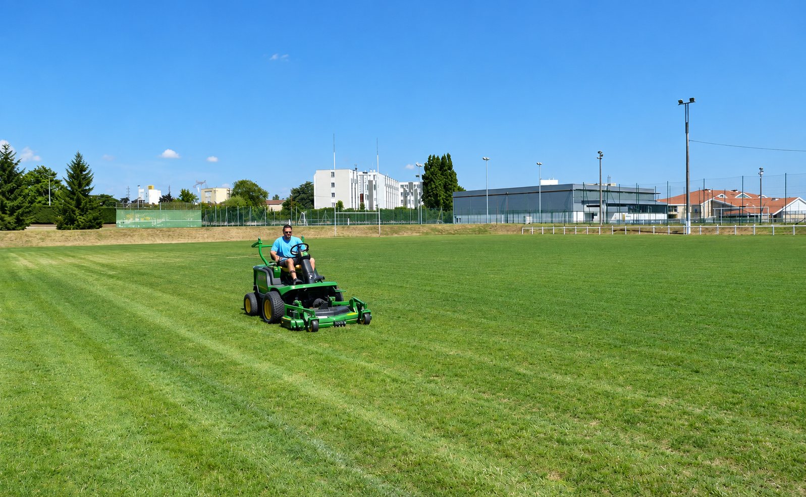 Un homme en t-shirt bleu conduit une tondeuse autoportée verte sur un grand terrain de football en herbe sous un ciel bleu.