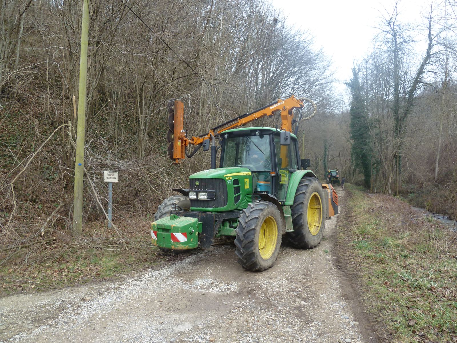 Un tracteur vert équipé d'un bras d'élagage orange travaille sur un chemin de gravier bordé d'arbres sans feuilles.