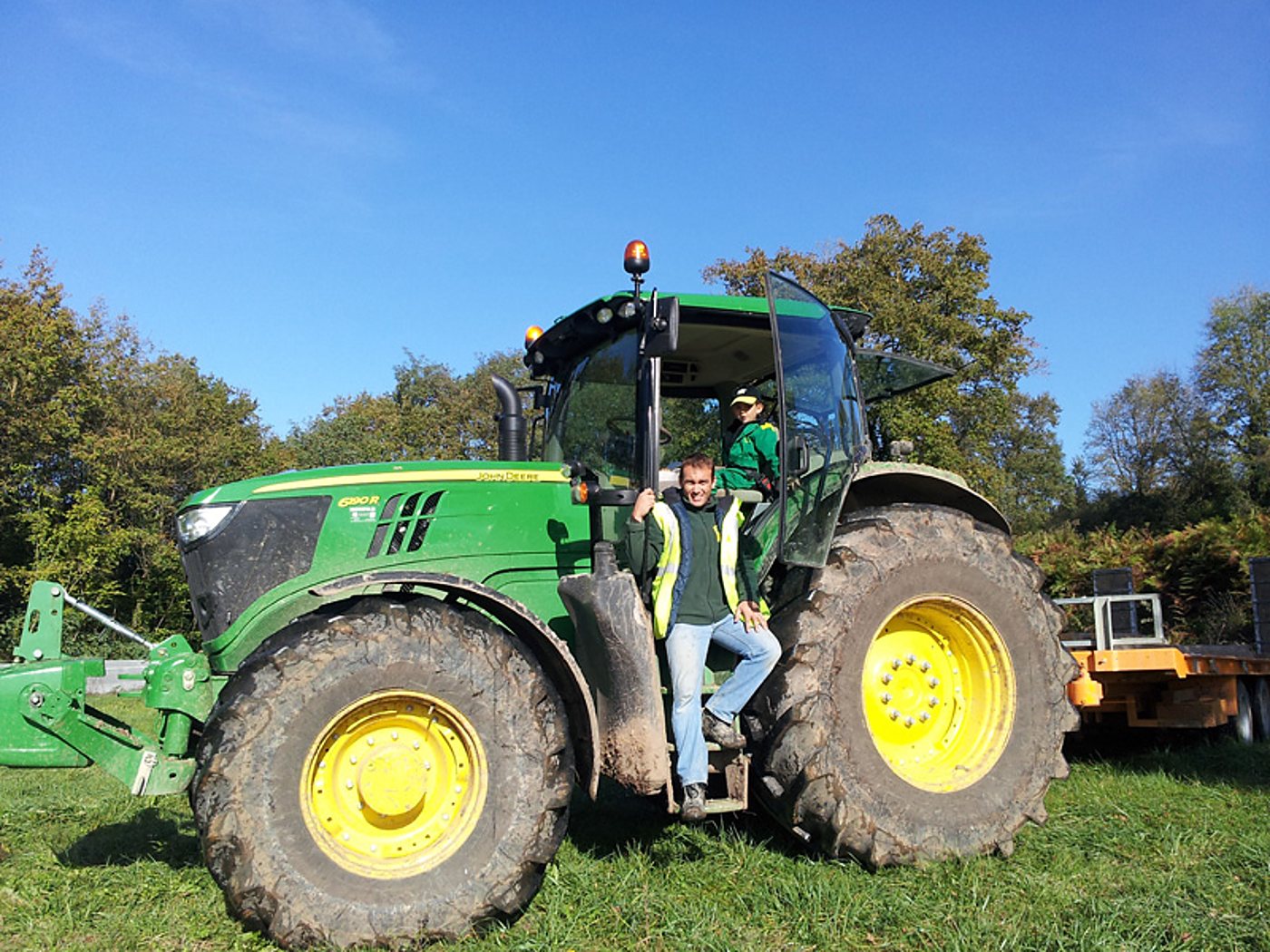 Un homme en gilet jaune se tient sur la marche d'un gros tracteur John Deere vert, tandis qu'un autre est assis dans la cabine, sur une prairie ensoleillée.
